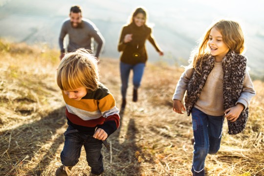 Smiling parents and two children walking or running together across dry brown grass.