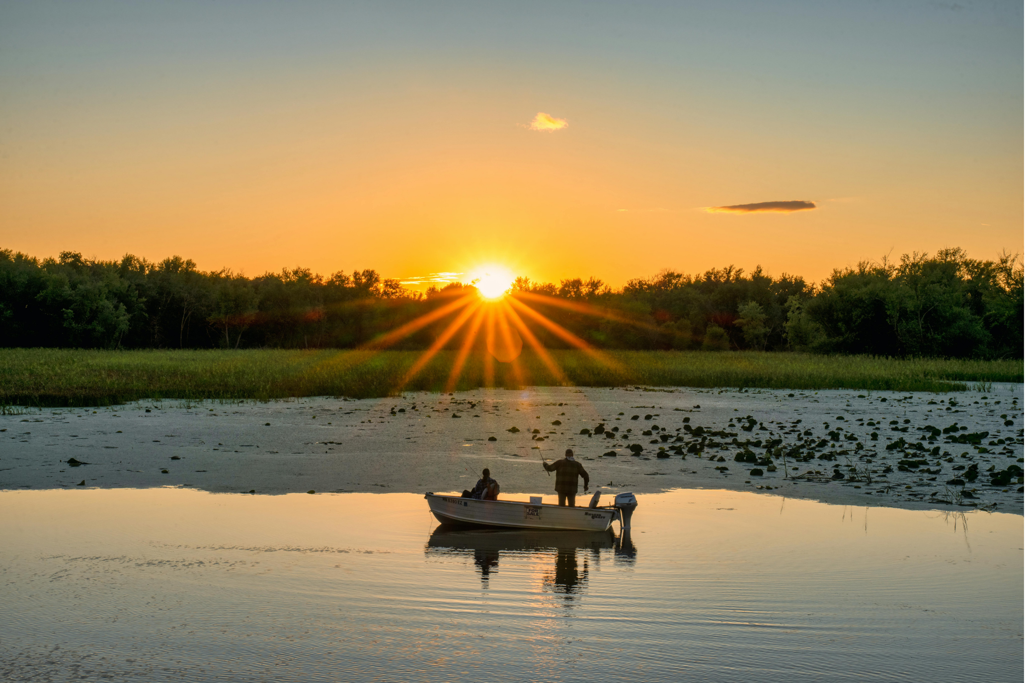 Picture of 2 people fishing in a boat on the water, with a background of trees and the sun setting over them